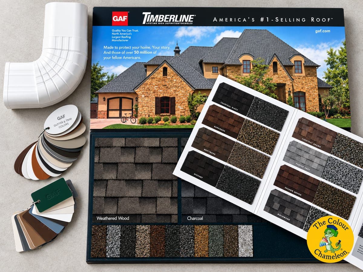 A GAF Timberline shingle sample board with an open swatch book of roof colors, a gutter and trim color fan, and a white downspout elbow laid out on a counter